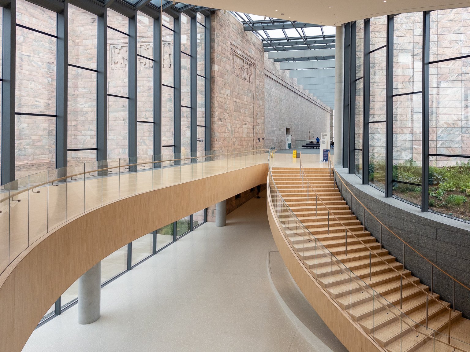 A curved wooden staircase with glass railings ascends within the modern interior of the Joslyn Art Museum. The space features large windows revealing stone walls and natural light from a skylight above.