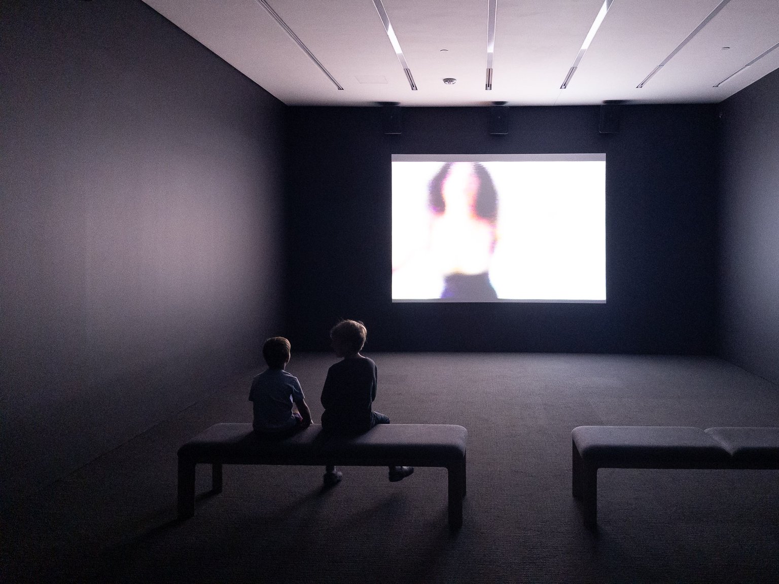 Two children sit on a bench in a dimly lit gallery, attentively watching a large, bright projection of an abstract image. The minimalist space features dark walls and modern lighting, emphasizing the immersive art experience.