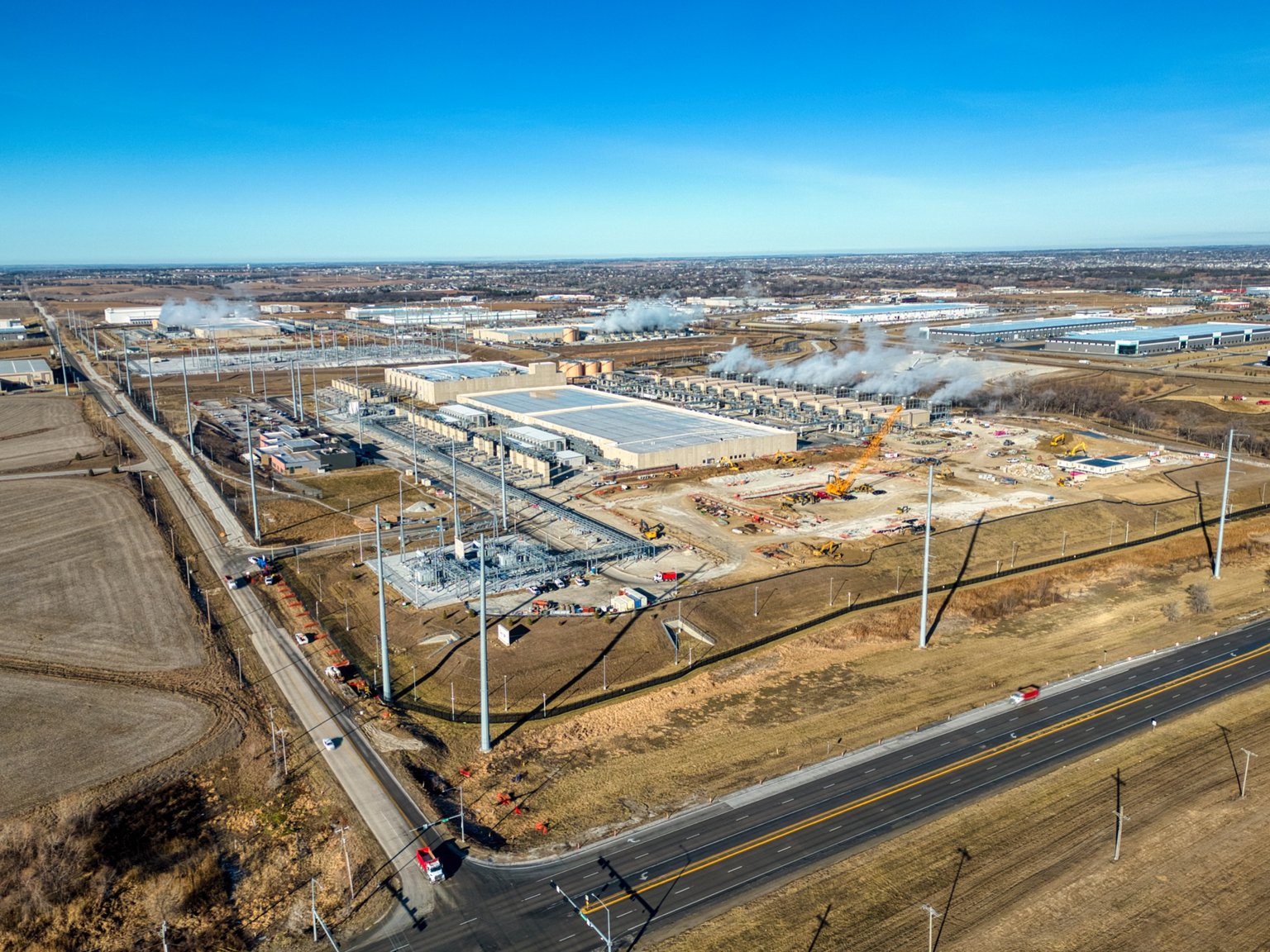 An aerial view captures a large-scale construction site for a Google data center in Omaha, Nebraska, with multiple buildings under development and heavy machinery active. The surrounding area includes roads, utility infrastructure, and open fields, all set against a clear blue sky.