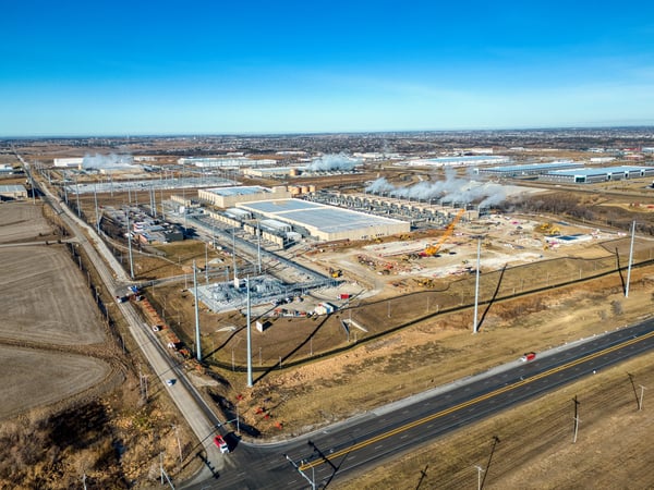 An aerial view captures a large-scale construction site for a Google data center in Omaha, Nebraska, with multiple buildings under development and heavy machinery active. The surrounding area includes roads, utility infrastructure, and open fields, all set against a clear blue sky.