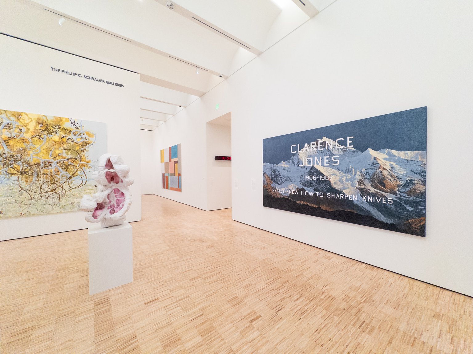 A modern art gallery features a large mountain-themed artwork with text honoring Clarence Jones, alongside other contemporary pieces including sculptures and abstract paintings. The space has white walls, light wooden flooring, and is illuminated by recessed ceiling lights.