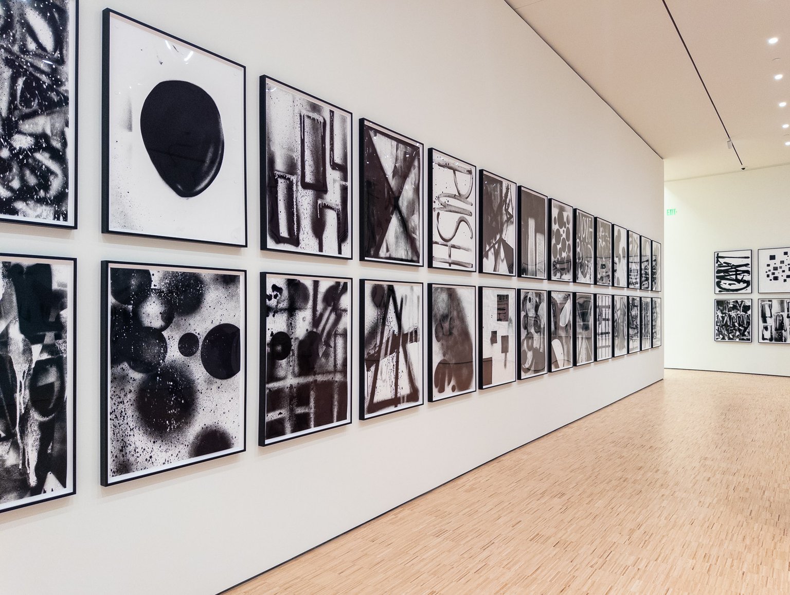 A long gallery wall features numerous framed black and white abstract artworks arranged in neat rows. The space has a minimalist design with light-colored walls, wooden flooring, and recessed lighting, creating an open, modern art museum environment.
